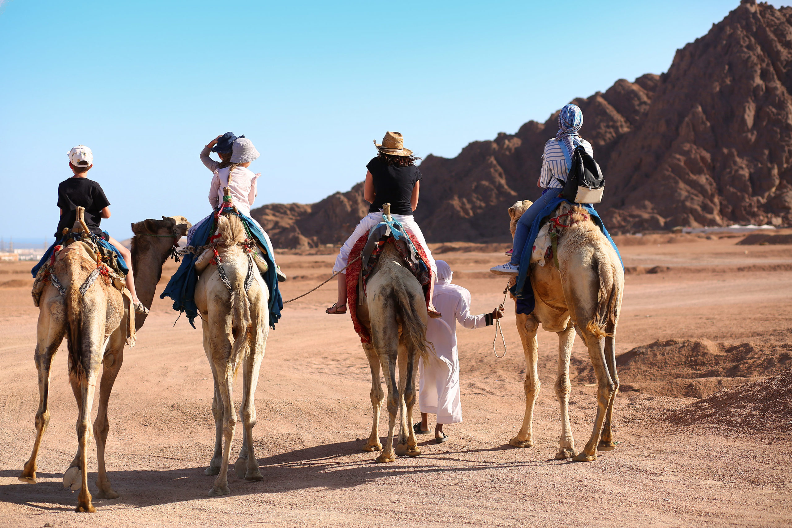Five people in casual clothing ride camels in the desert, being led by a guide.