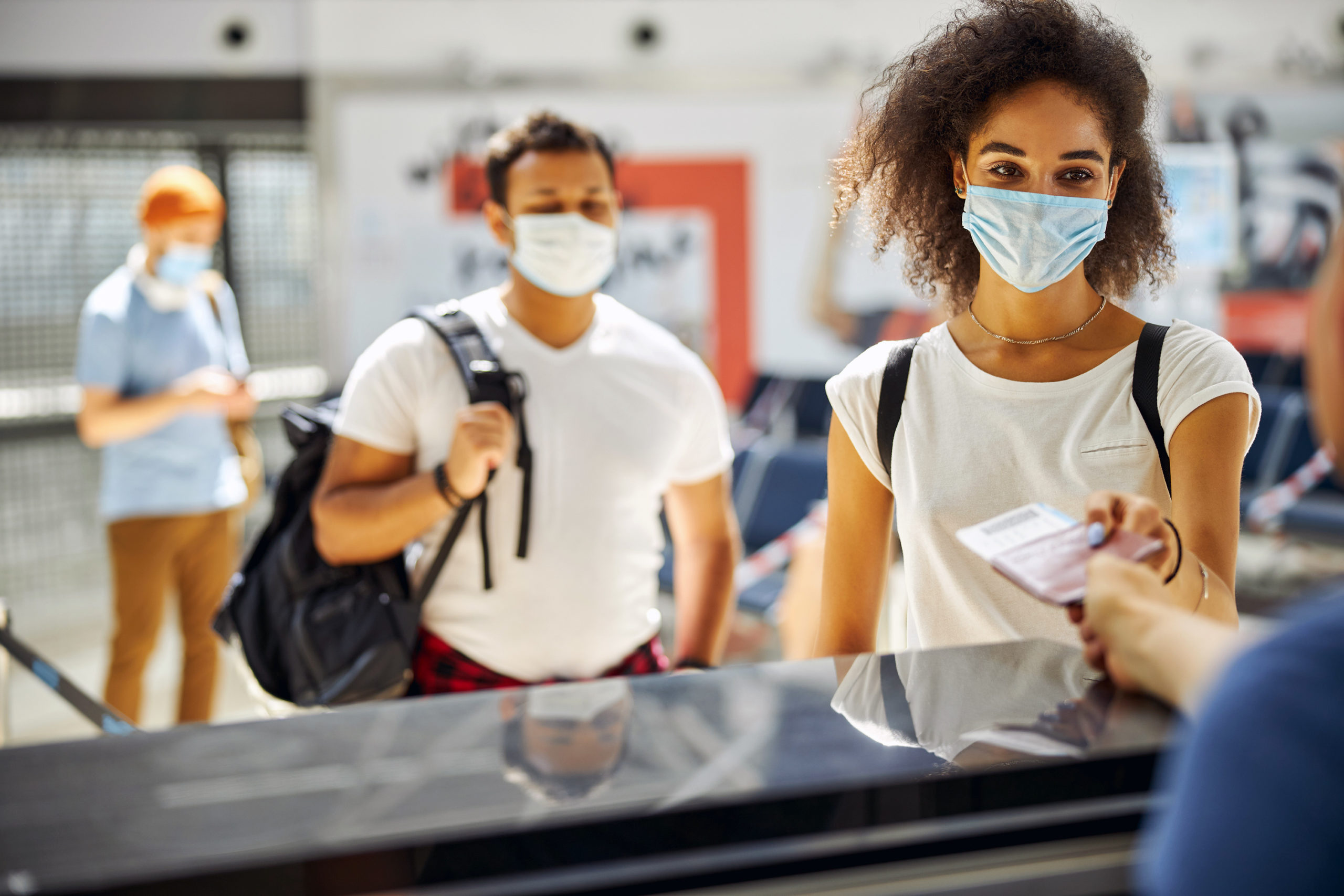 A masked woman with a backpack handing information to a worker behind a counter, with two other masked people wait in line behind her social distancing.