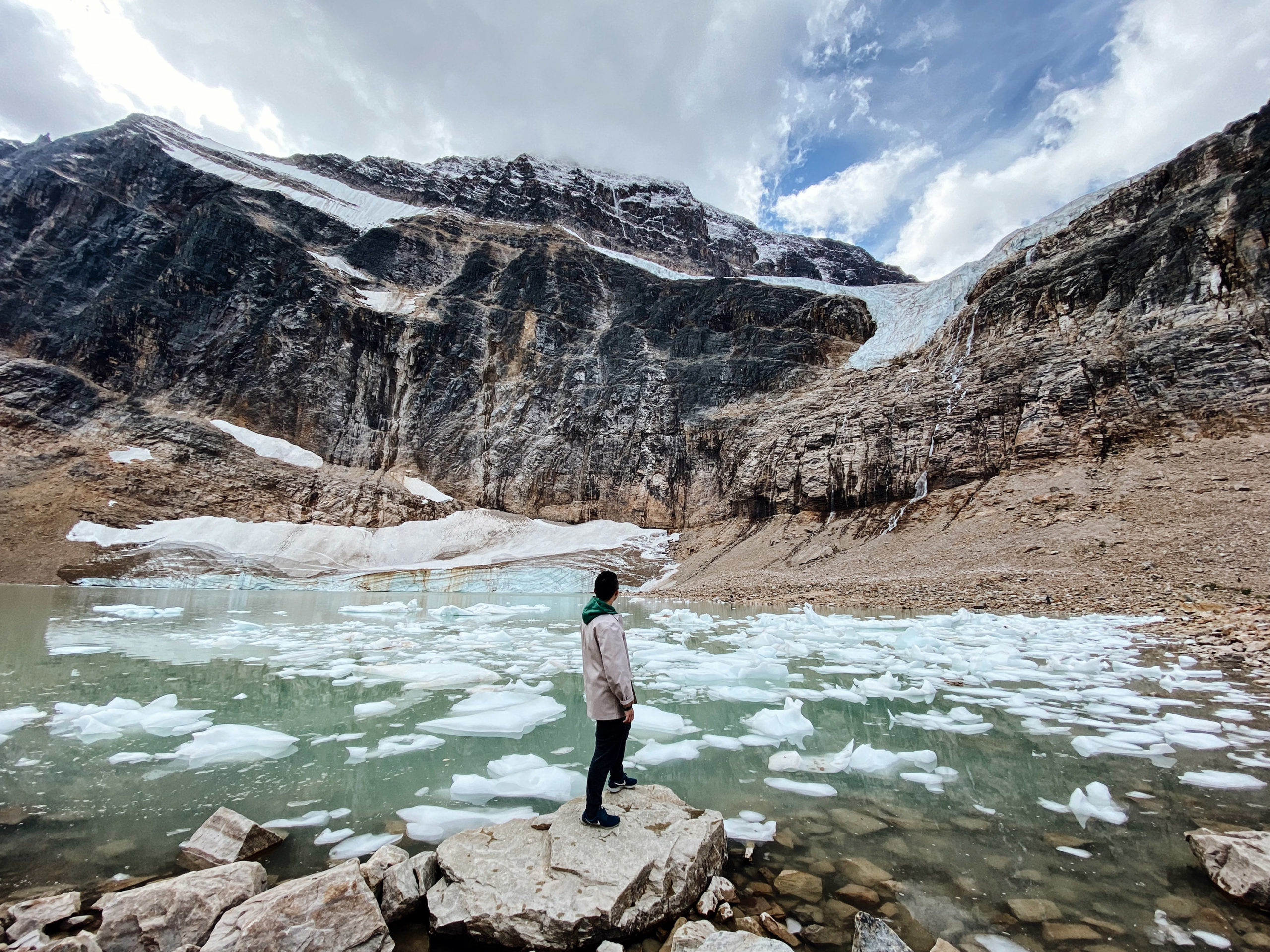 A man dressed warmly in layers stands on rocks overlooking water with large ice chucks and a rugged, snow-capped mountain scene.