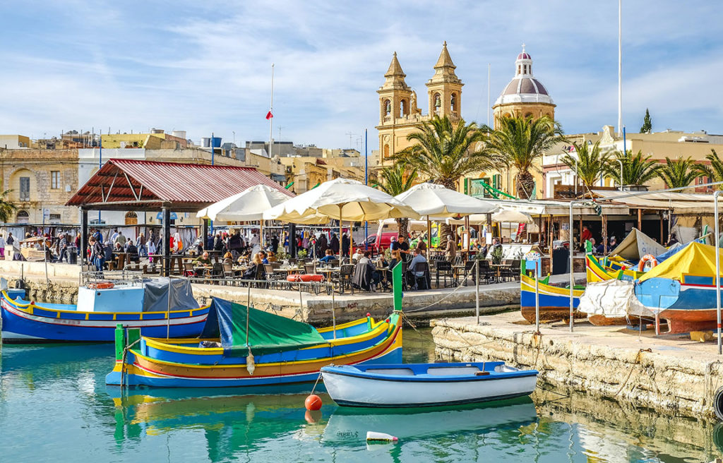 The bustling island of Malta, one of Sky Bird Travel & Tours top travel destinations 2023, colorful boats along the port city with travelers at a market.
