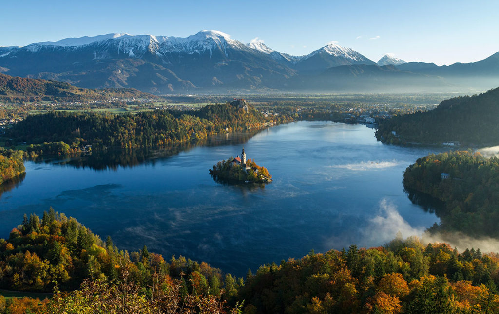 A panorama of Slovenia's lakes, forests, and mountains with a solo island with a lighthouse in this vacation spot by Sky Bird Travel & Tours top travel destinations of 2023.
